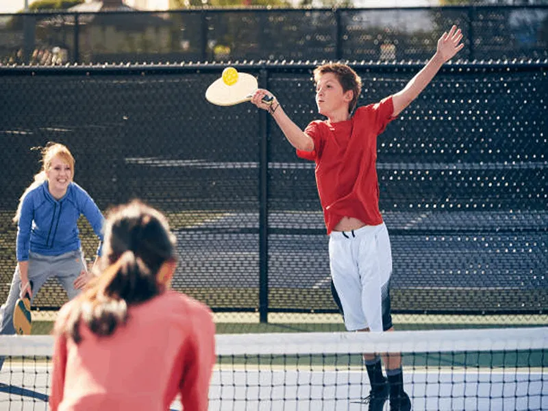 Boy in a red shirt hitting the wiffle ball with a pickleball paddle, illustrating Pickleball for Kids.