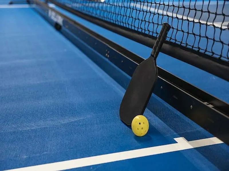 an image of a pickleball paddle placed on the floor with a yellow wiffle ball beside it.
